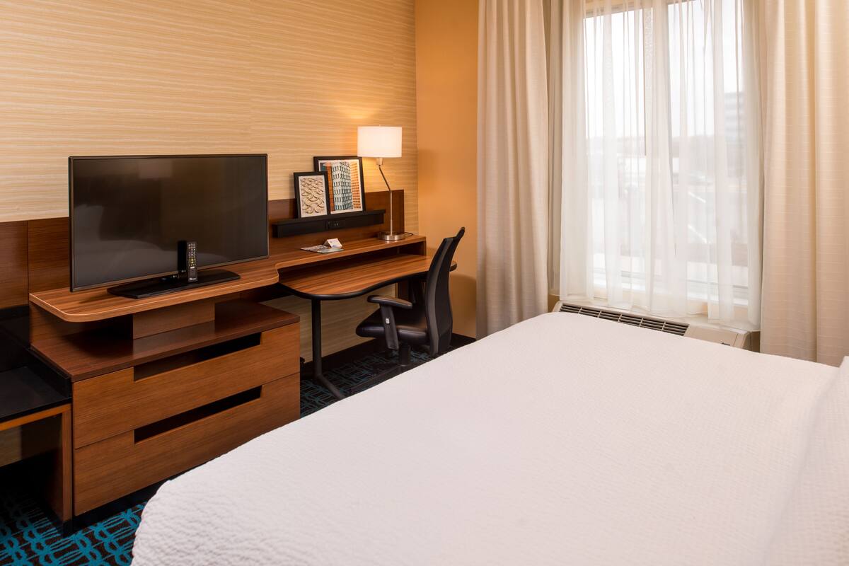 A hotel room with a bed, a desk with a TV, a chair, a lamp, and a window with curtains.