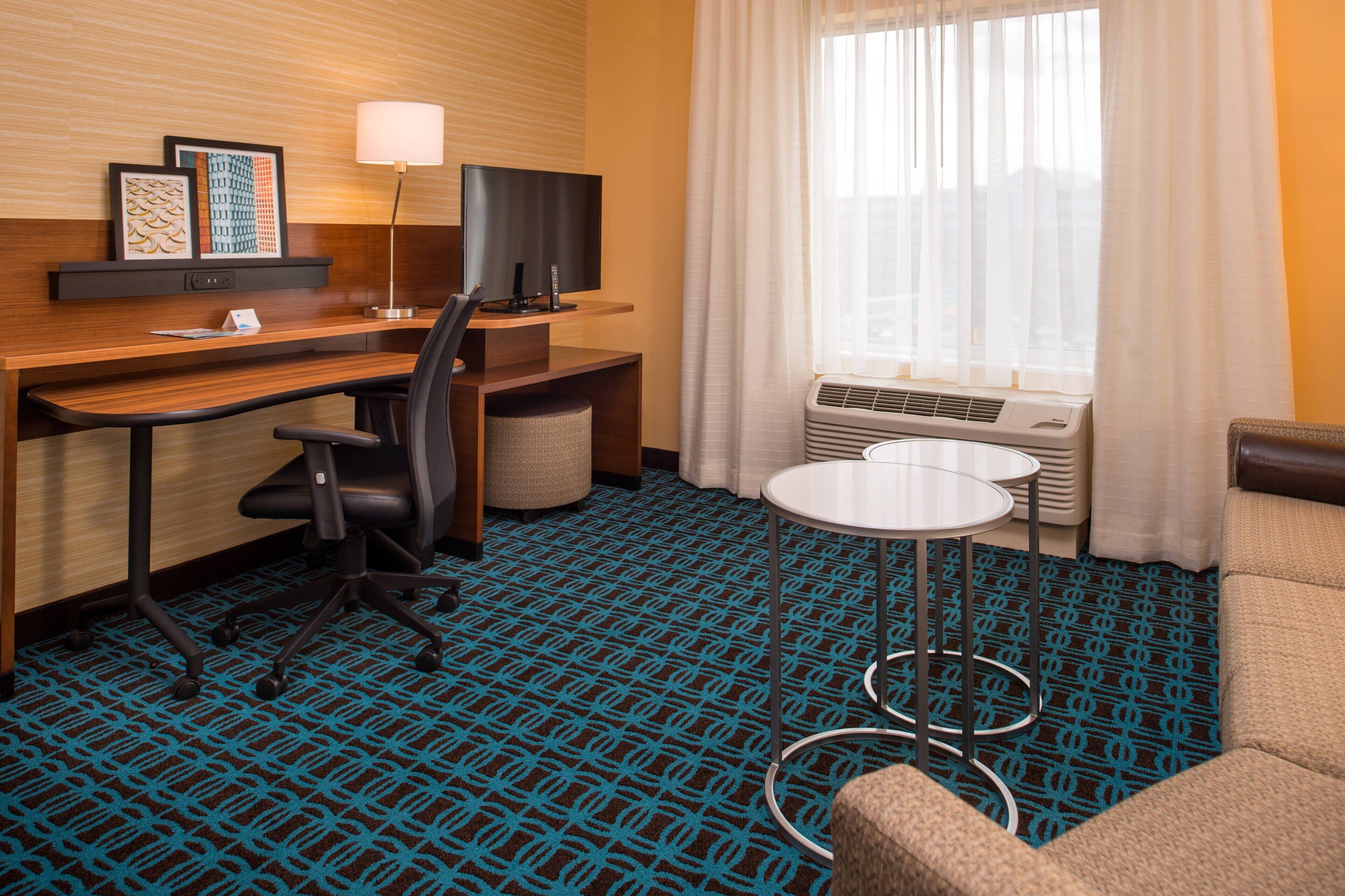 A hotel room with a desk, chair, TV, lamp, and a small round table.