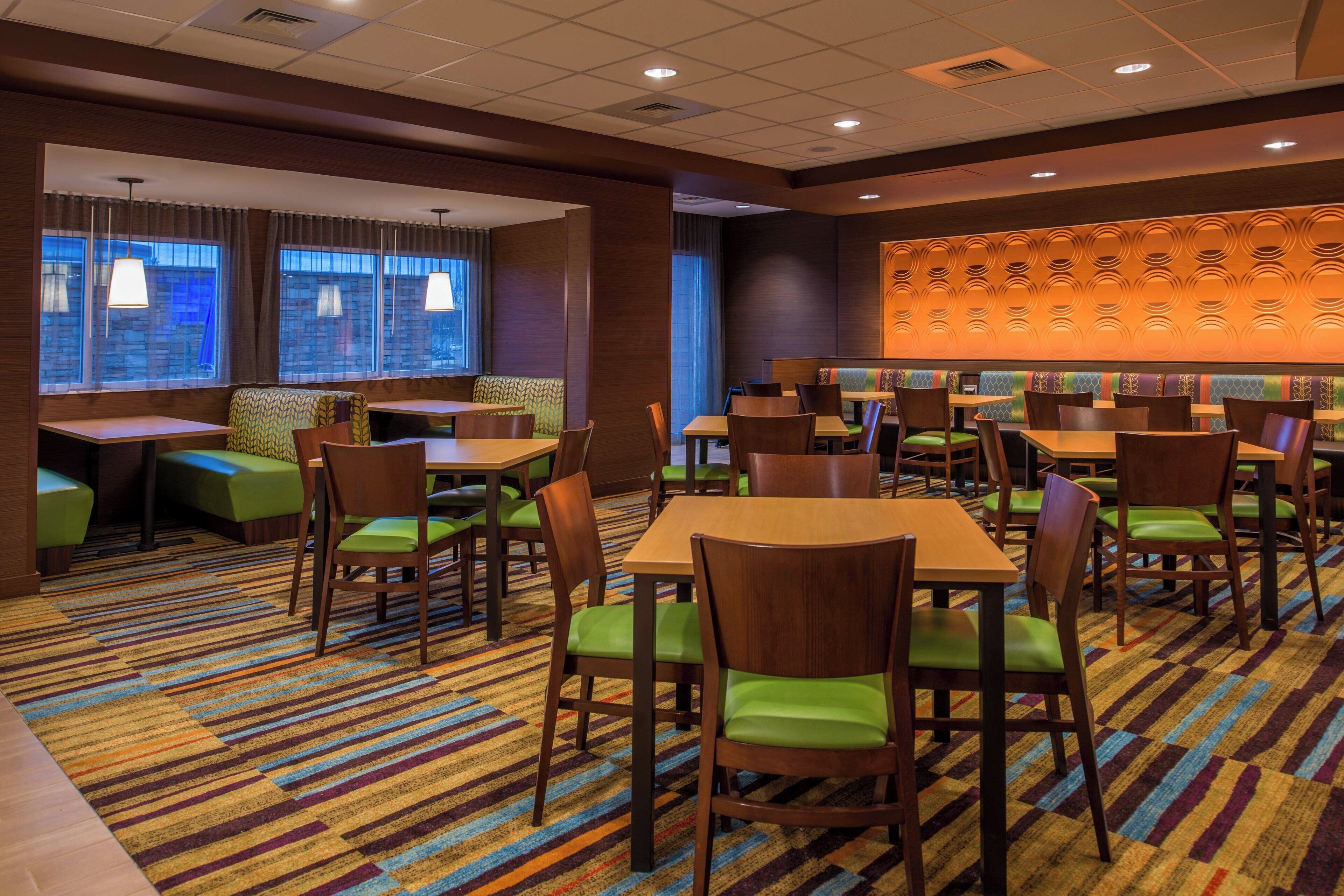 A hotel dining area with multiple tables and chairs, a patterned carpet, and a wall with a decorative panel.