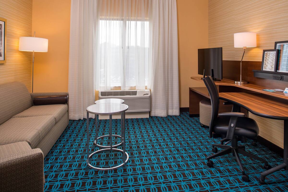 A hotel room with a sofa, a desk with a chair, a small round table, a TV, and a window with curtains.