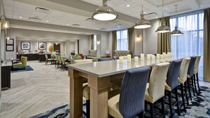 A hotel dining area with a long table, multiple chairs, pendant lighting, and a kitchen in the background. - Hampton Inn Livonia Detroit (Livonia)