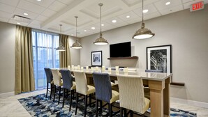 A conference room with a long table, chairs, pendant lights, a TV, framed pictures, and an exit sign. - Hampton Inn Livonia Detroit (Livonia)