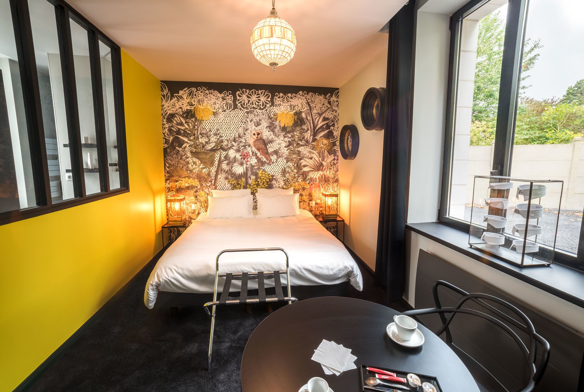 Ein Hotelzimmer mit Bett, kleinem Tisch mit Tasse und Untertasse, Stuhl, Fenster mit Blick nach draußen und einer Wand mit floralem Dekor.
