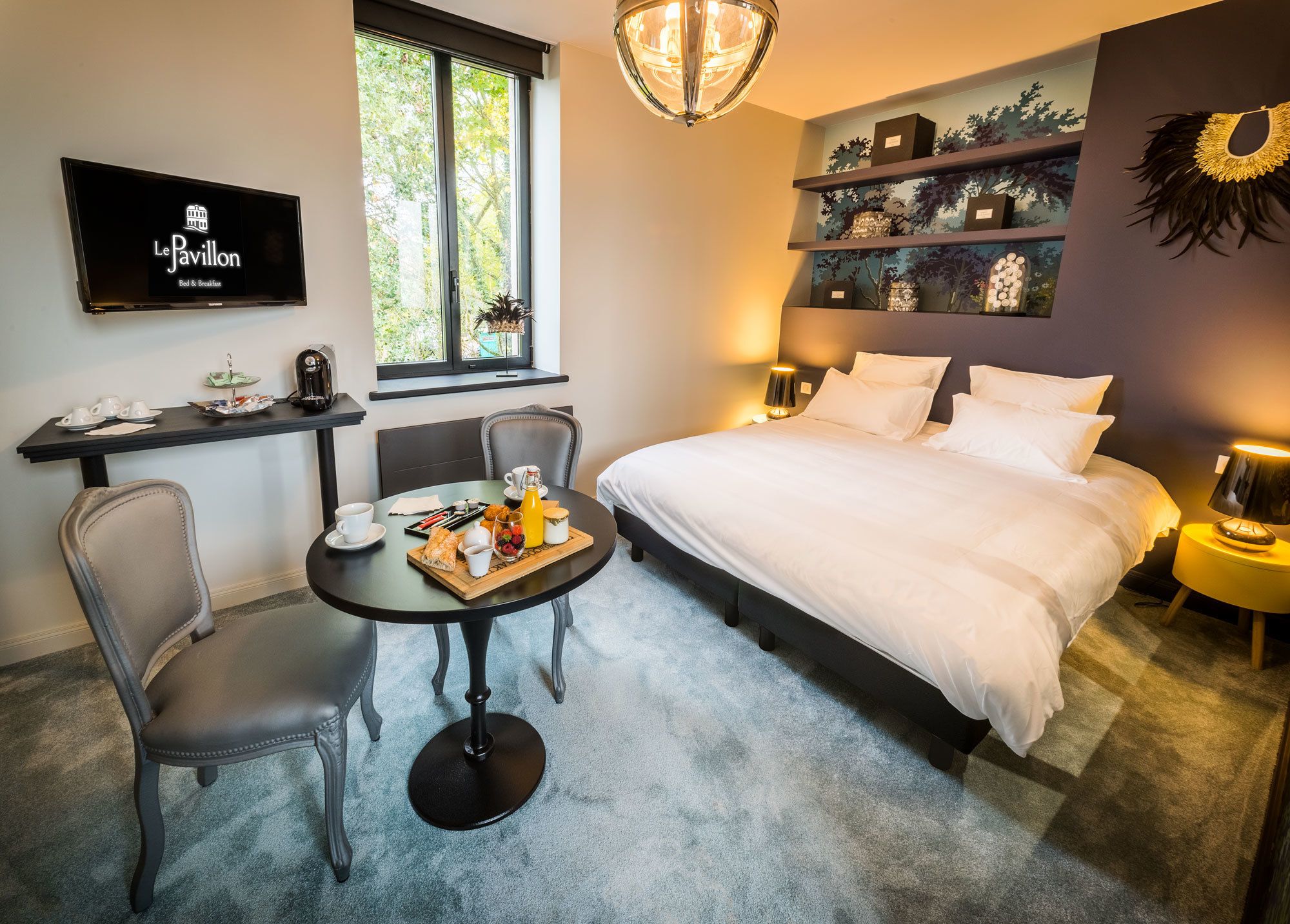 Une chambre d’hôtel comprenant un lit, un bureau avec des articles pour le petit-déjeuner, une télévision et une fenêtre donnant sur des arbres.