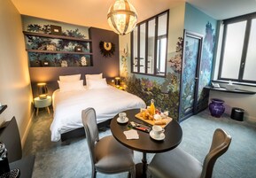 Habitación de hotel con cama, una mesa redonda con alimentos para el desayuno, un escritorio con ordenador y un ventanal grande.
