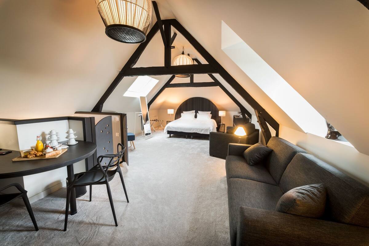 A cozy attic bedroom with a bed, a sofa, a dining table with chairs, and a chandelier.
