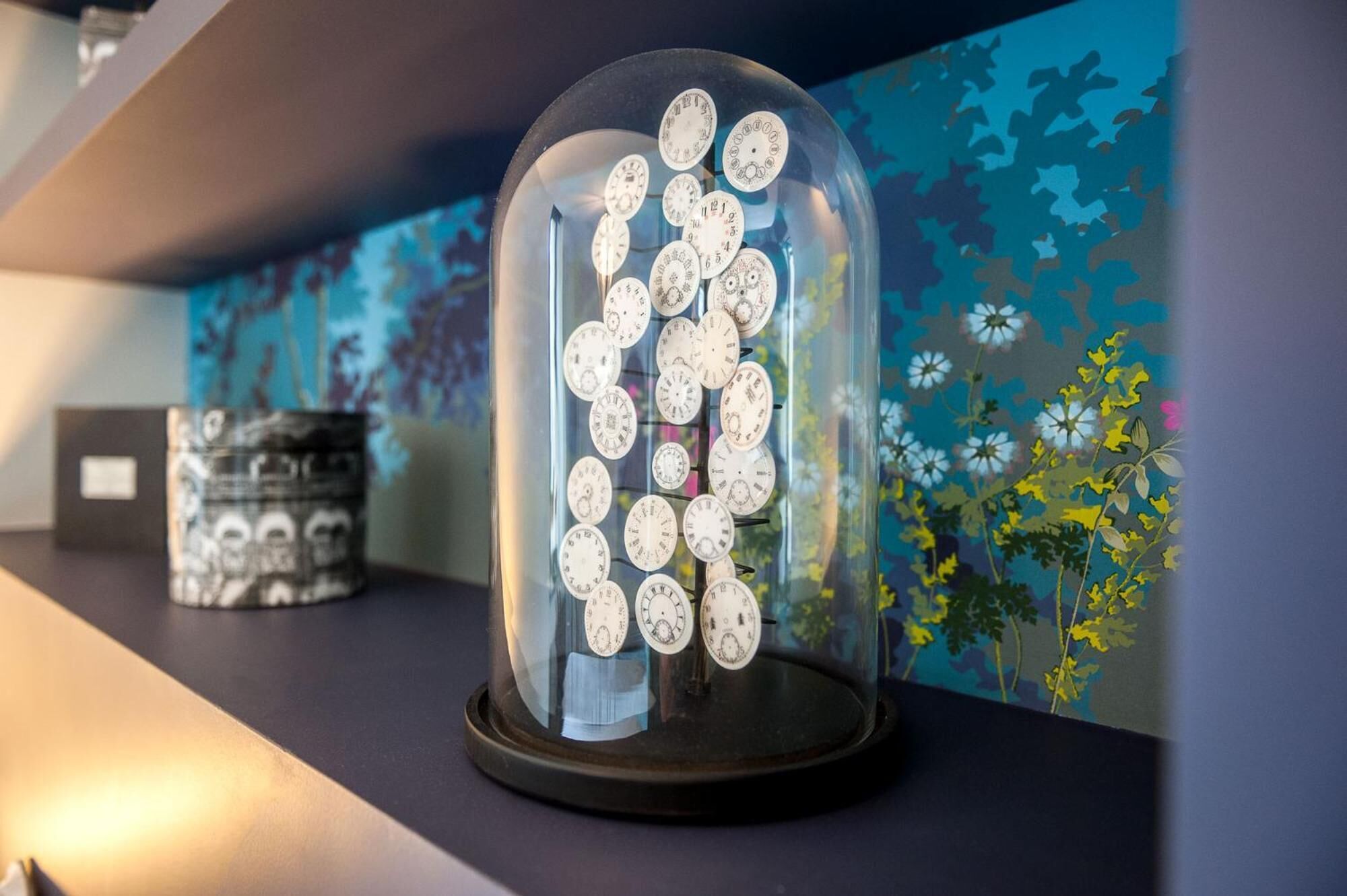 A glass dome display case containing vintage clocks on a dark surface with a floral wallpaper background.