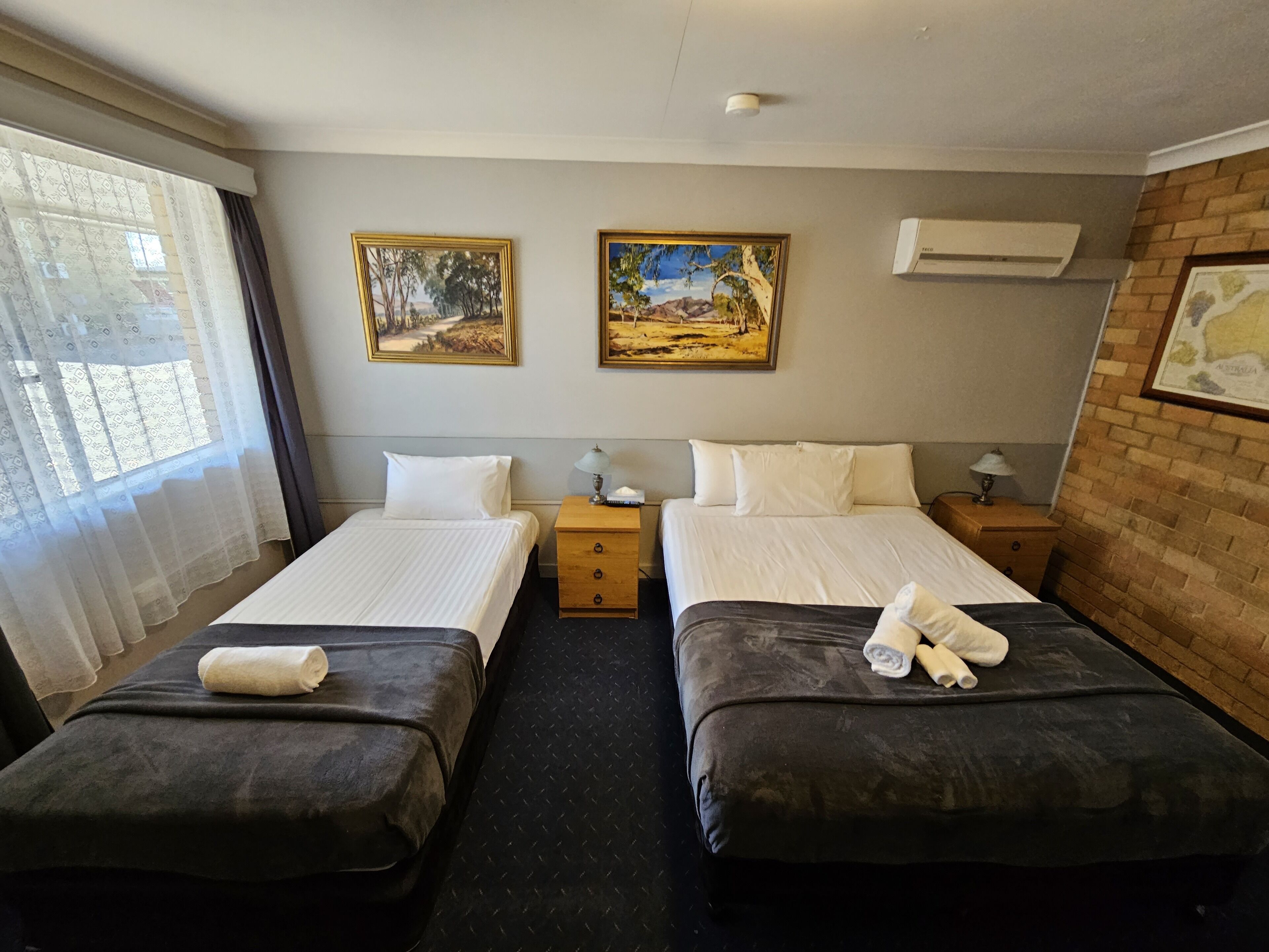 Ein Hotelzimmer mit zwei Betten, einem Vorhangfenster, zwei Bildern an der Wand und einer Klimaanlage.