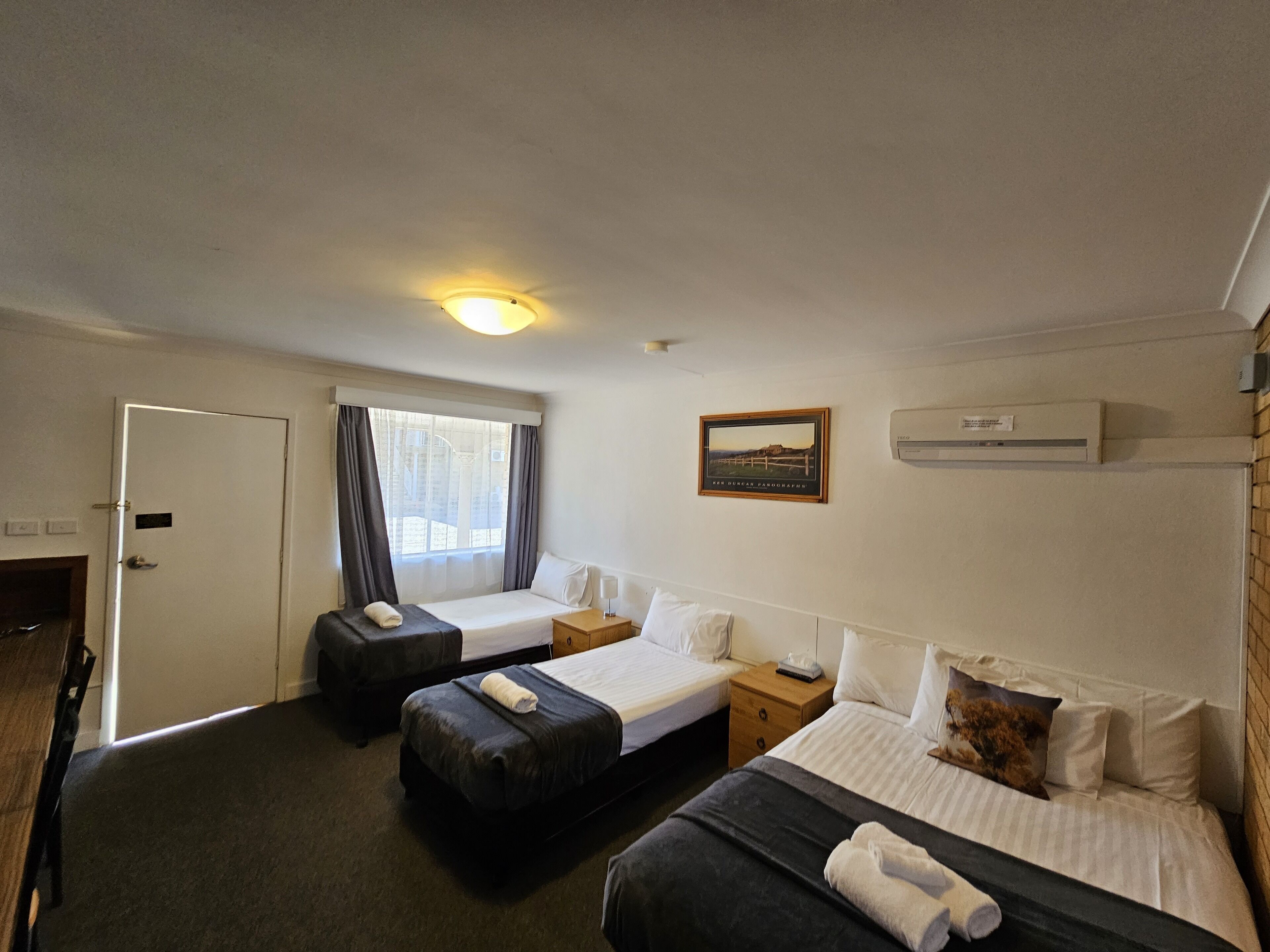 A hotel room with two beds, a desk, a chair, a window with curtains, a wall-mounted air conditioner, and a framed picture.