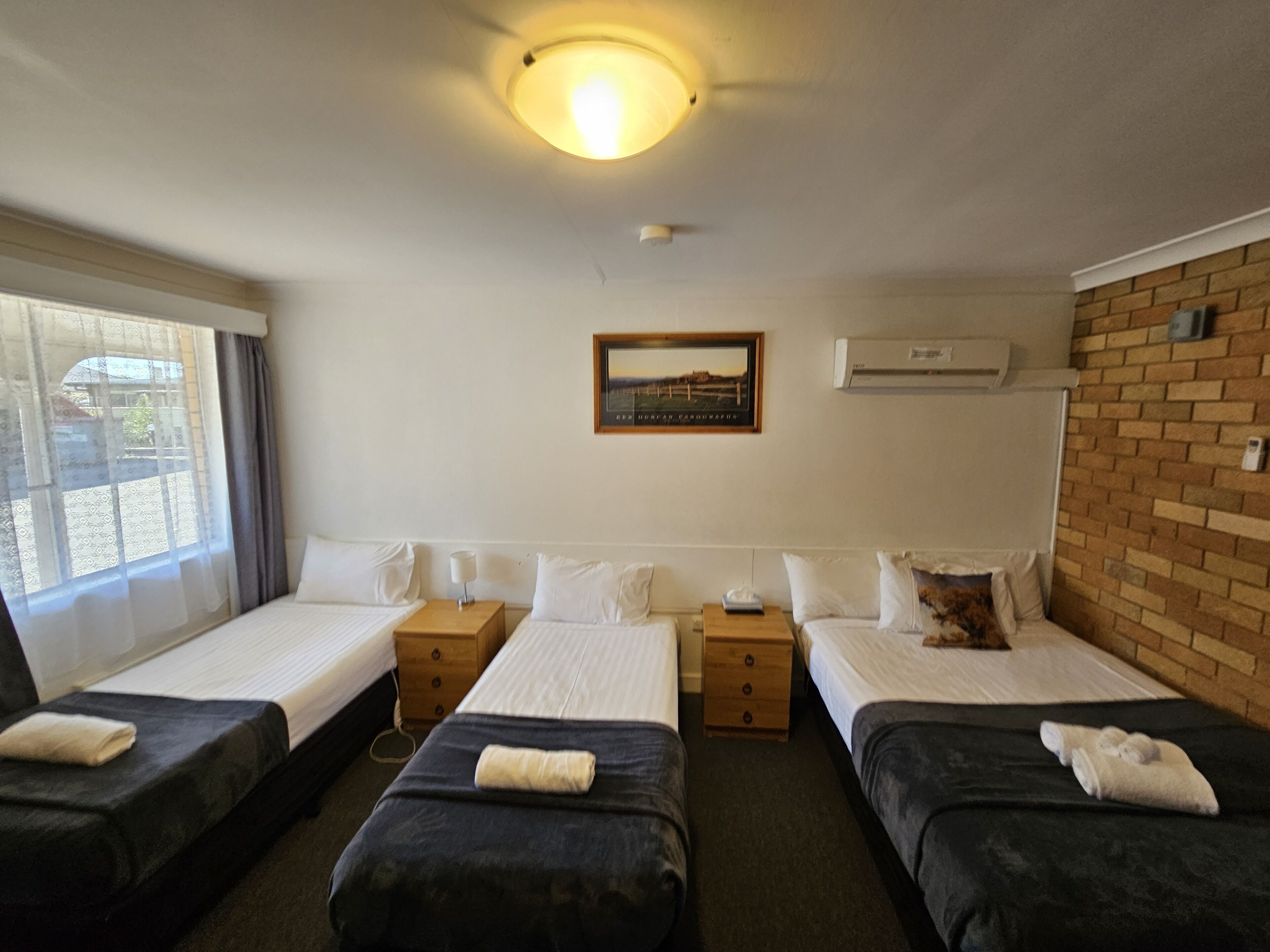 Habitación de hotel con dos camas individuales, una mesita, una mesita de noche, aire acondicionado en la pared y una ventana con cortinas.