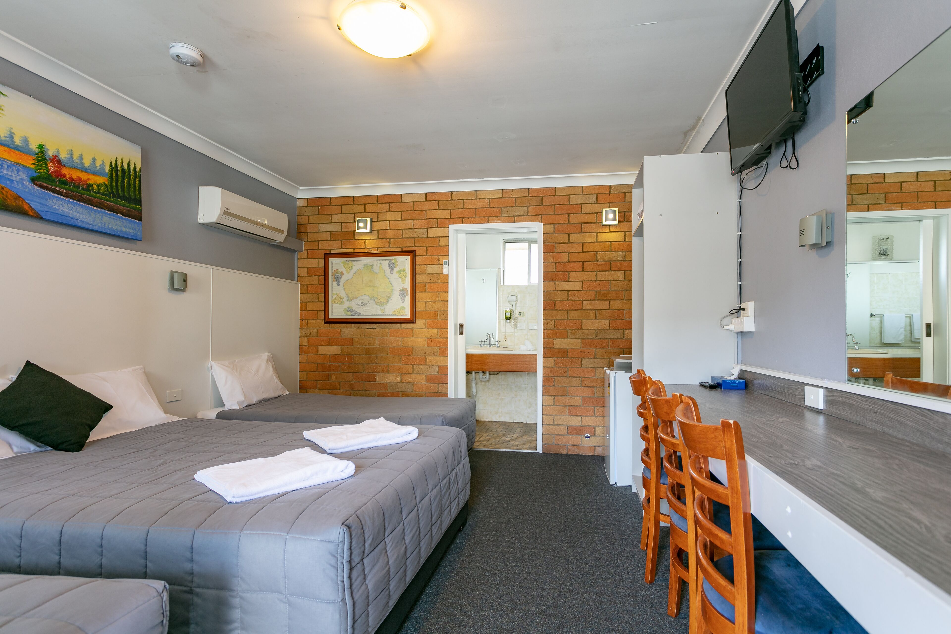 A hotel room with two beds, a desk with chairs, a television, and a bathroom visible through an open door.