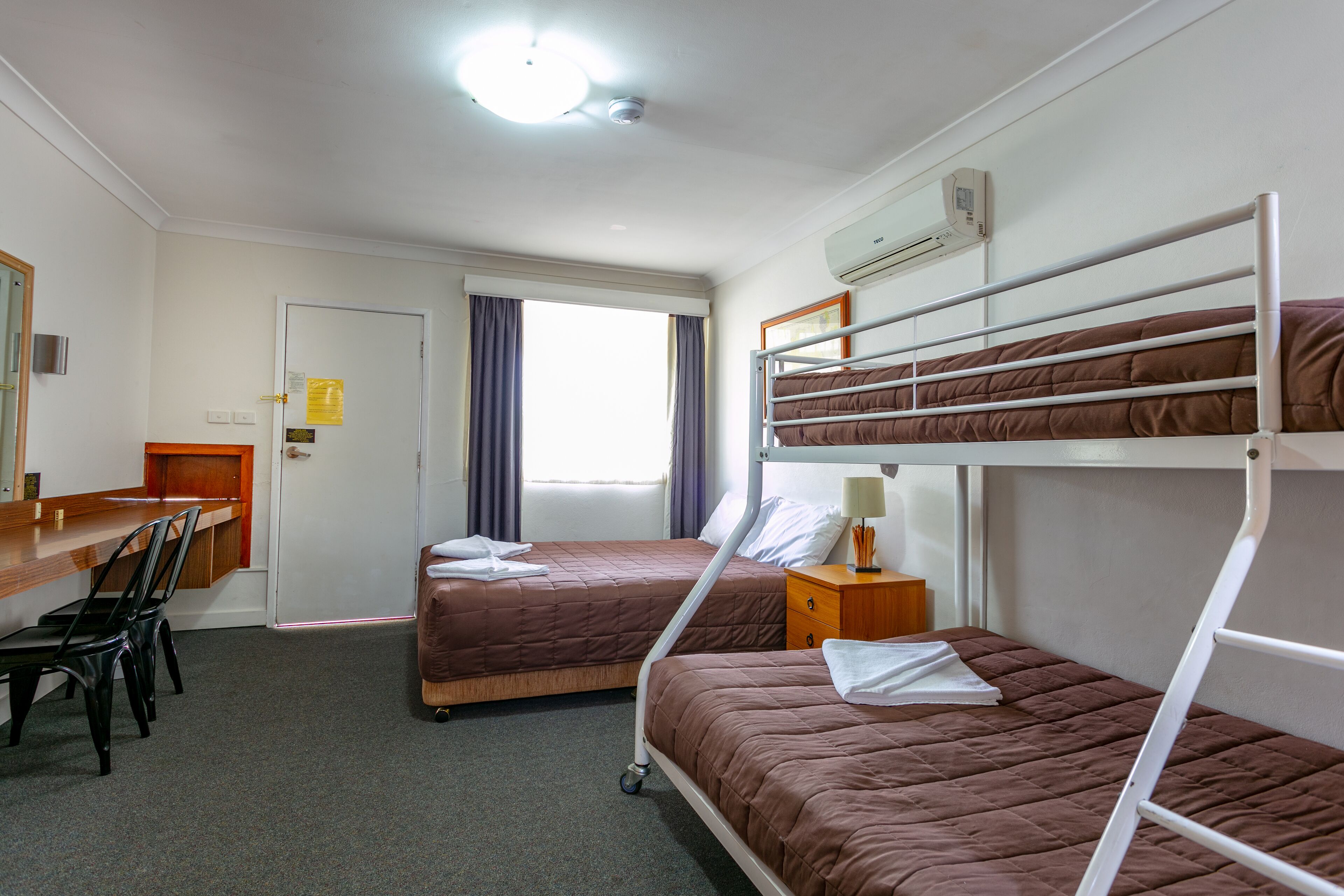 Habitació d'hotel amb lliteres, una taula de treball amb cadires, una petita taula de nit i una finestra amb cortines.