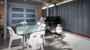An outdoor patio with a table and chairs, a grill, and a covered area with a garage door in the background. - Hunter Valley Motel (Cessnock)