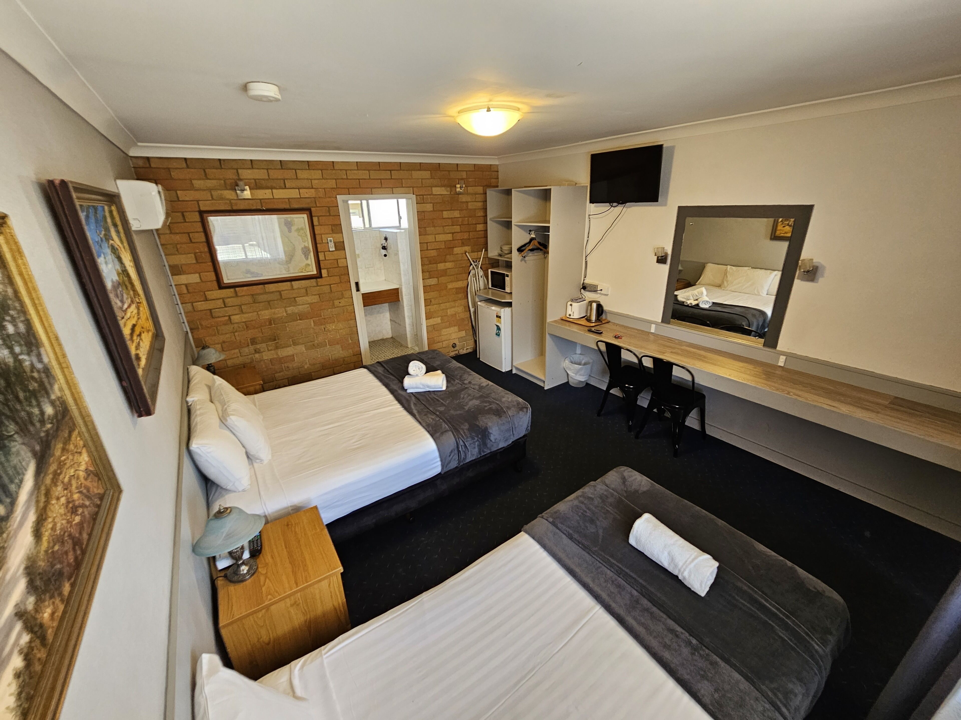 Habitación de hotel con dos camas, un escritorio, un espejo, un televisor y una pared de ladrillo.