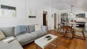 Un salon moderne avec un canapé gris, une table basse et un coin repas avec des chaises en bois. On aperçoit une cuisine à l’arrière-plan, avec un lave-linge et un four à micro-ondes.
