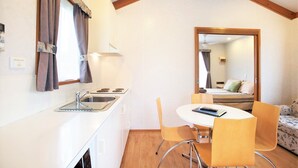 A compact kitchen with a sink, stove, and dining area with chairs. A mirror reflects a bedroom with a bed and a window.