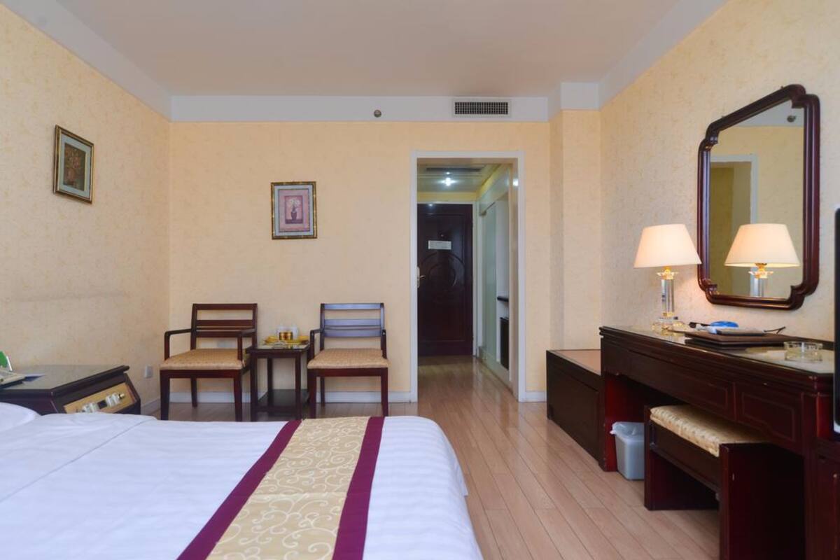 Ein Hotelzimmer mit einem großen Bett, zwei Stühlen, einem Schreibtisch, einem Schminktisch mit Spiegel und einer Mülleimer.