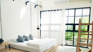 A modern bedroom with a wooden platform bed, white bedding, and a view of greenery through large windows. - The Companion - Hostel (Bangkok)