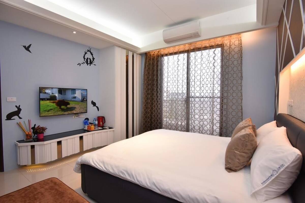 A modern hotel room with a large bed, a flat-screen TV mounted on the wall, a window with patterned curtains, and a ceiling fan.