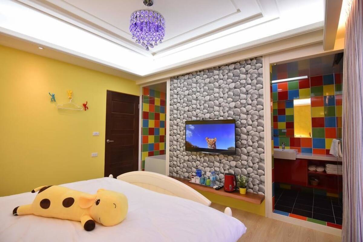 A bedroom with a bed, a television mounted on a stone wall, a colorful shelf, and a plush toy on the bed.
