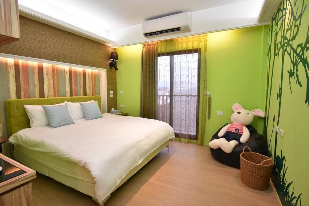 A bedroom with a large bed, a nightstand, a wall-mounted lamp, a window with curtains, a green wall with a mural, and a bean bag chair with a stuffed toy.