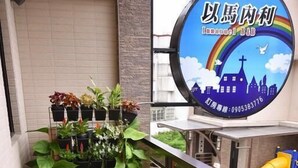 Un balcon avec une table en verre et des chaises blanches, un arrangement de plantes en pot et un panneau représentant un arc-en-ciel et la silhouette d’une église.