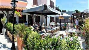 Un café en plein air, avec des tables et des chaises, entouré de verdure et bordé d’un bâtiment blanc aux volets bruns.