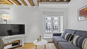Un salón con televisor de pantalla plana, un sofá, una mesita redonda y una ventana con vistas a edificios.