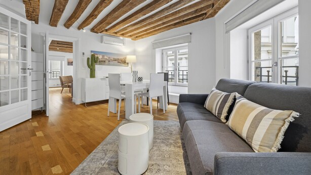 Una sala de estar moderna con un sofá gris, vigas de madera en el techo y un comedor con mobiliario blanco.