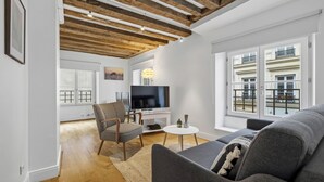 Una sala de estar moderna con vigas de madera a la vista, un televisor de pantalla plana, un sofá gris y vistas a un edificio con balcones.