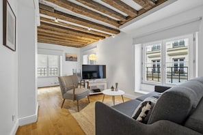 A modern living room with exposed wooden beams, a flat-screen TV, a grey sofa, and a view of a building with balconies.