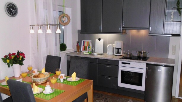 A compact kitchen with modern appliances, a dining table set for breakfast, and a clock on the wall. - Self-in Goslar, suitor Apartment - Erdgescschoss (Goslar)
