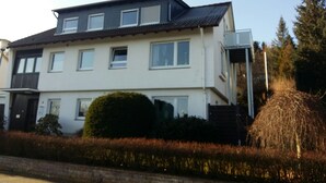 A two-story residential building with a flat roof, multiple windows, and a small garden area with a hedge and a tree. - Self-in Goslar, suitor Apartment - Erdgescschoss (Goslar)