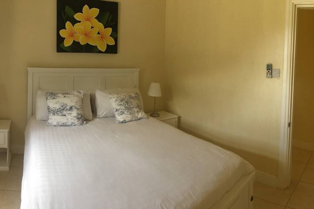 A neatly made bed with white bedding and two patterned pillows, a bedside table with a lamp, and a painting of yellow flowers on the wall.