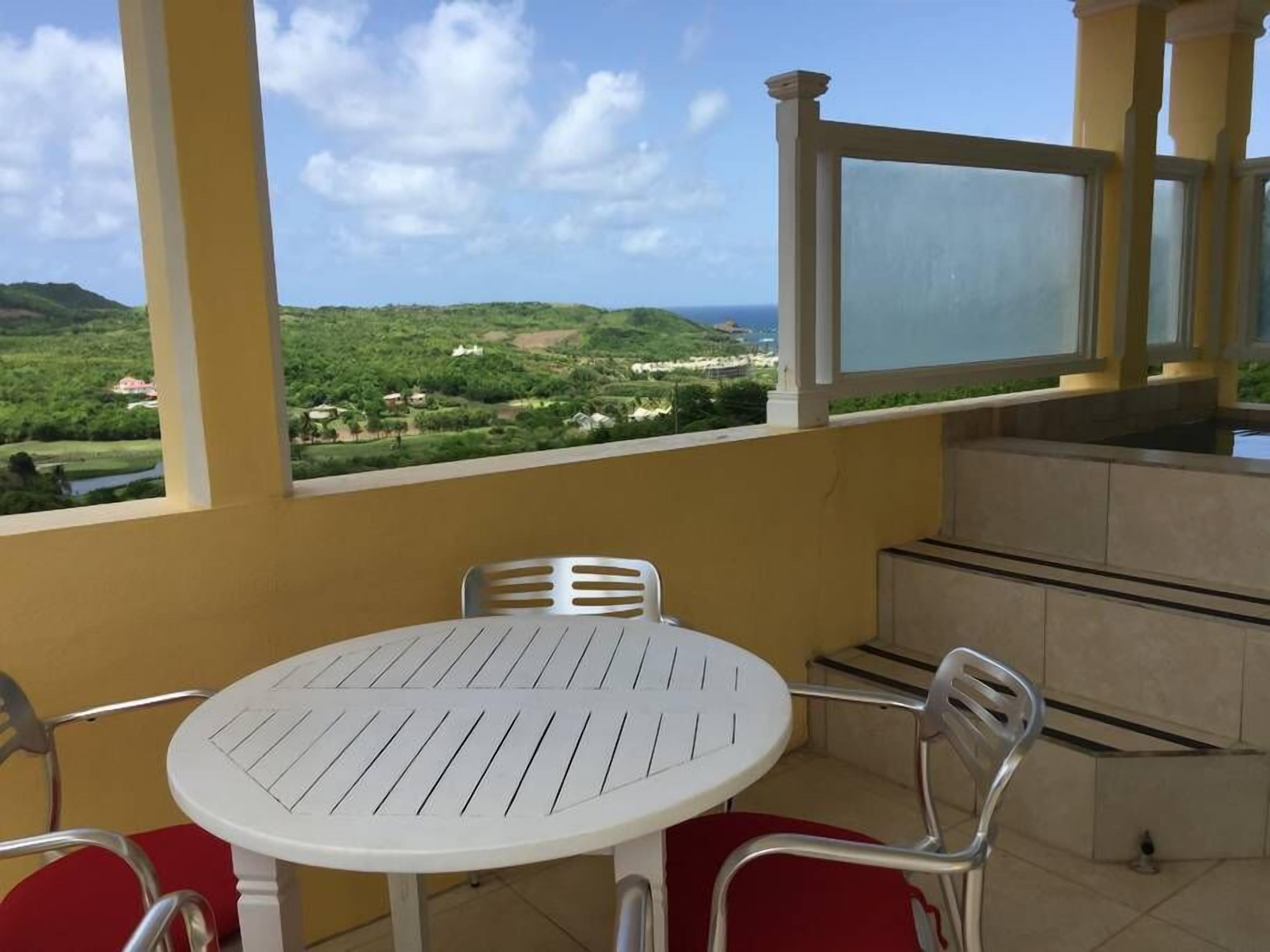 Un balcon avec une table et des chaises blanches, donnant sur un paysage verdoyant et une étendue d’eau.