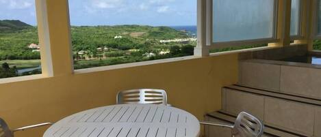 Un balcon avec une table et des chaises blanches, donnant sur un paysage verdoyant et une étendue d’eau.