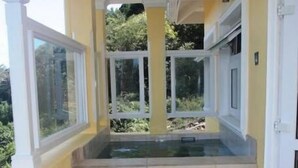 A sunlit balcony with a hot tub, a chair, and a view of greenery.