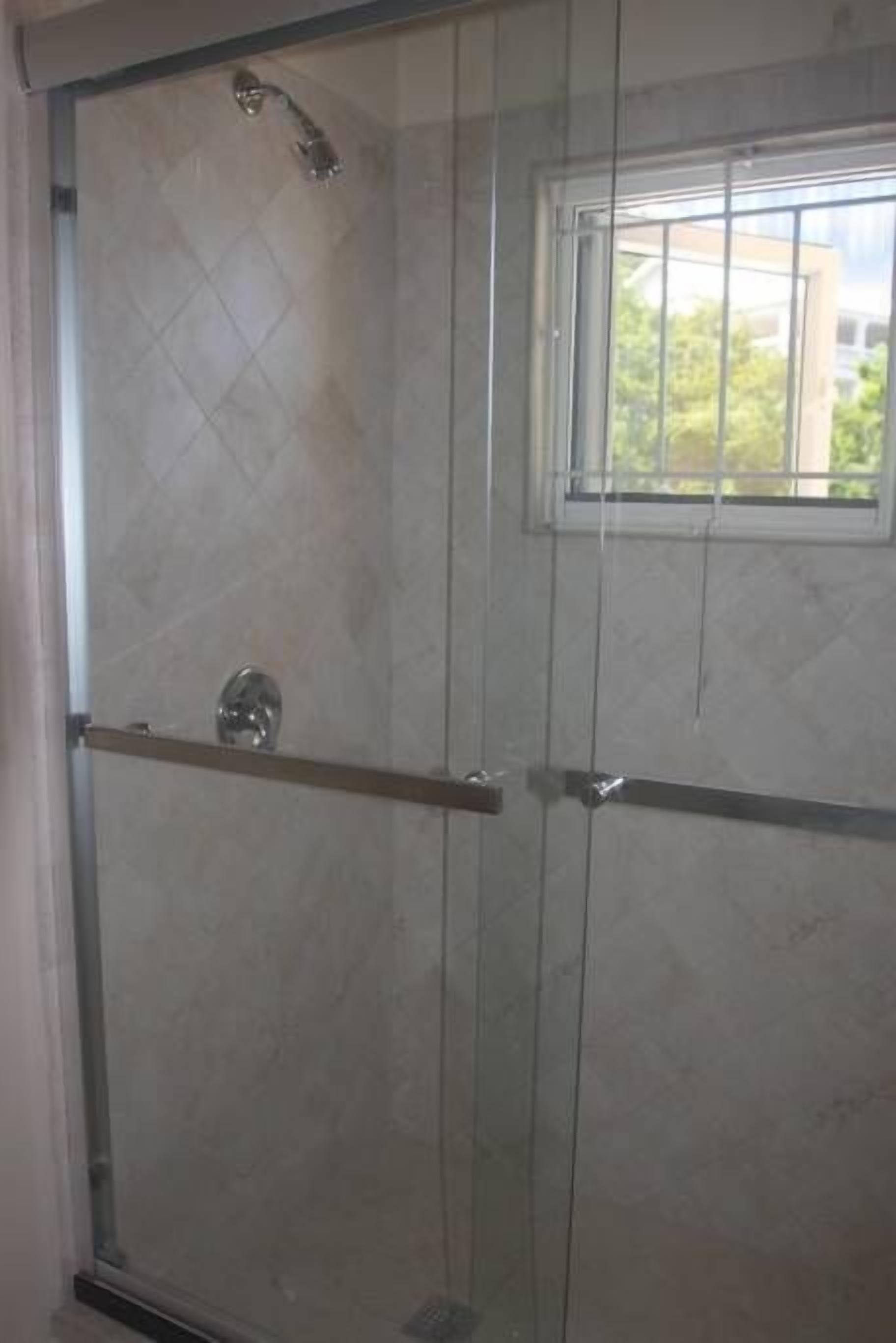 Une douche fermée par des parois de verre, dotée d’une fenêtre, d’une poignée en métal et d’un sol carrelé.
