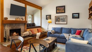 A cozy living room with a fireplace, a blue sofa, a wooden coffee table, and a TV mounted on the wall. - Outpost: Geranium 2622 (Wilson)
