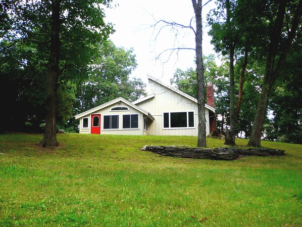 A single-story house with a red door, white siding, and a chimney. It is surrounded by trees and grassy area.