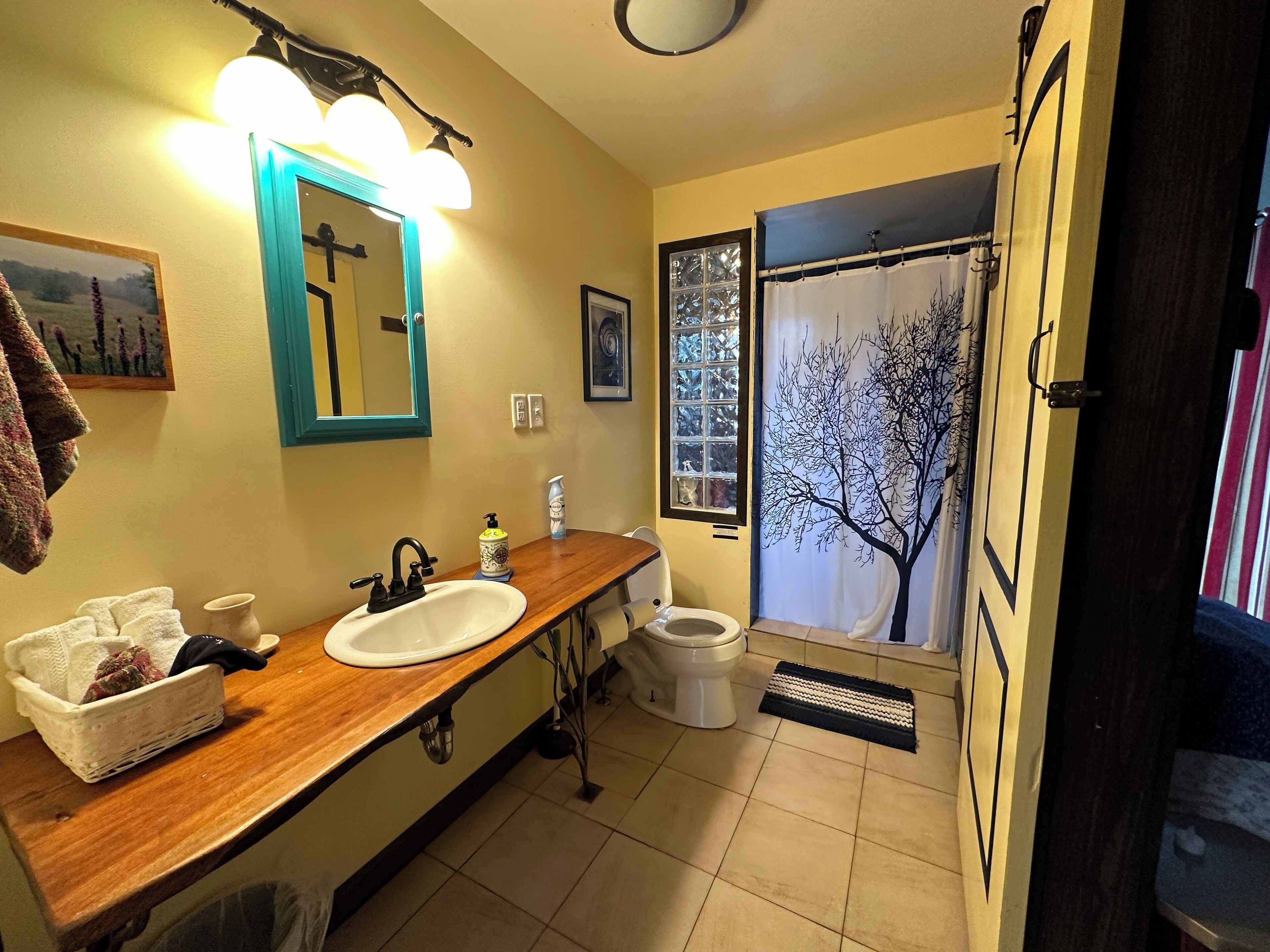A bathroom with a wooden vanity, a white sink, a mirror with lights, a window with a tree view, and a shower curtain with a tree design.