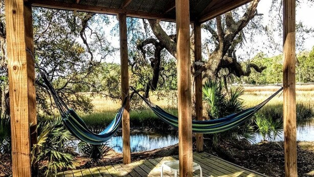 Une terrasse en bois avec un hamac et une chaise, entourée d’arbres et bordée par un plan d’eau.