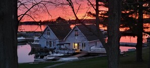 A waterfront property with a two-story house, docked boats, and a serene sunset view.
