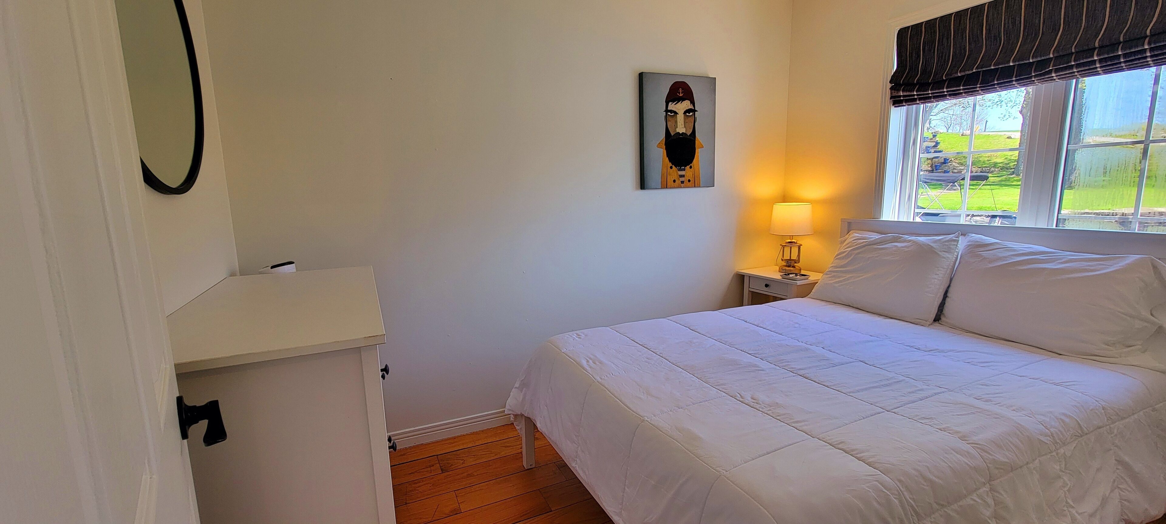 Un dormitorio con cama, mesita de noche, lámpara, un cuadro y ventana con vistas a vegetación.