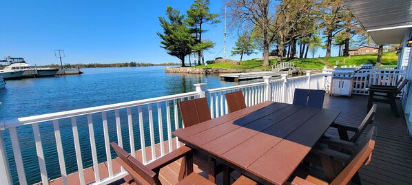 Eine Terrasse mit Tisch und Stühlen mit Blick auf einen ruhigen See, in der Nähe liegen Boote angedockt und es gibt eine Rasenfläche mit Bäumen.