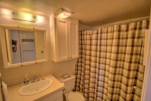 A bathroom with a white sink, mirror, and toilet. There is a plaid shower curtain and a cabinet above the toilet.