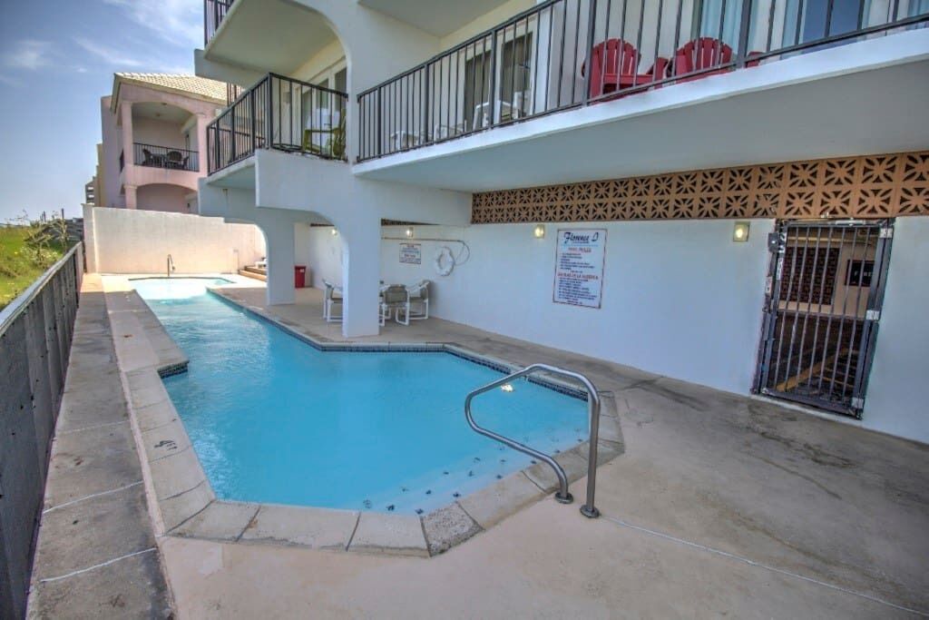A swimming pool with a metal ladder, a covered seating area, and a multi-level building with balconies.