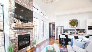 A spacious living area with a brick fireplace, a flat-screen TV, and a modern kitchen with white cabinetry.
