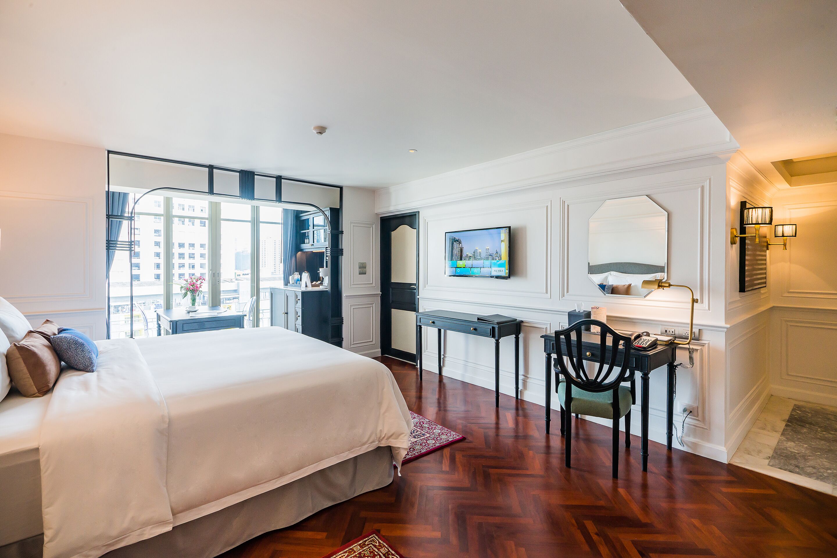 A hotel room with a bed, a desk, a chair, a TV, and a view of the city through large windows.