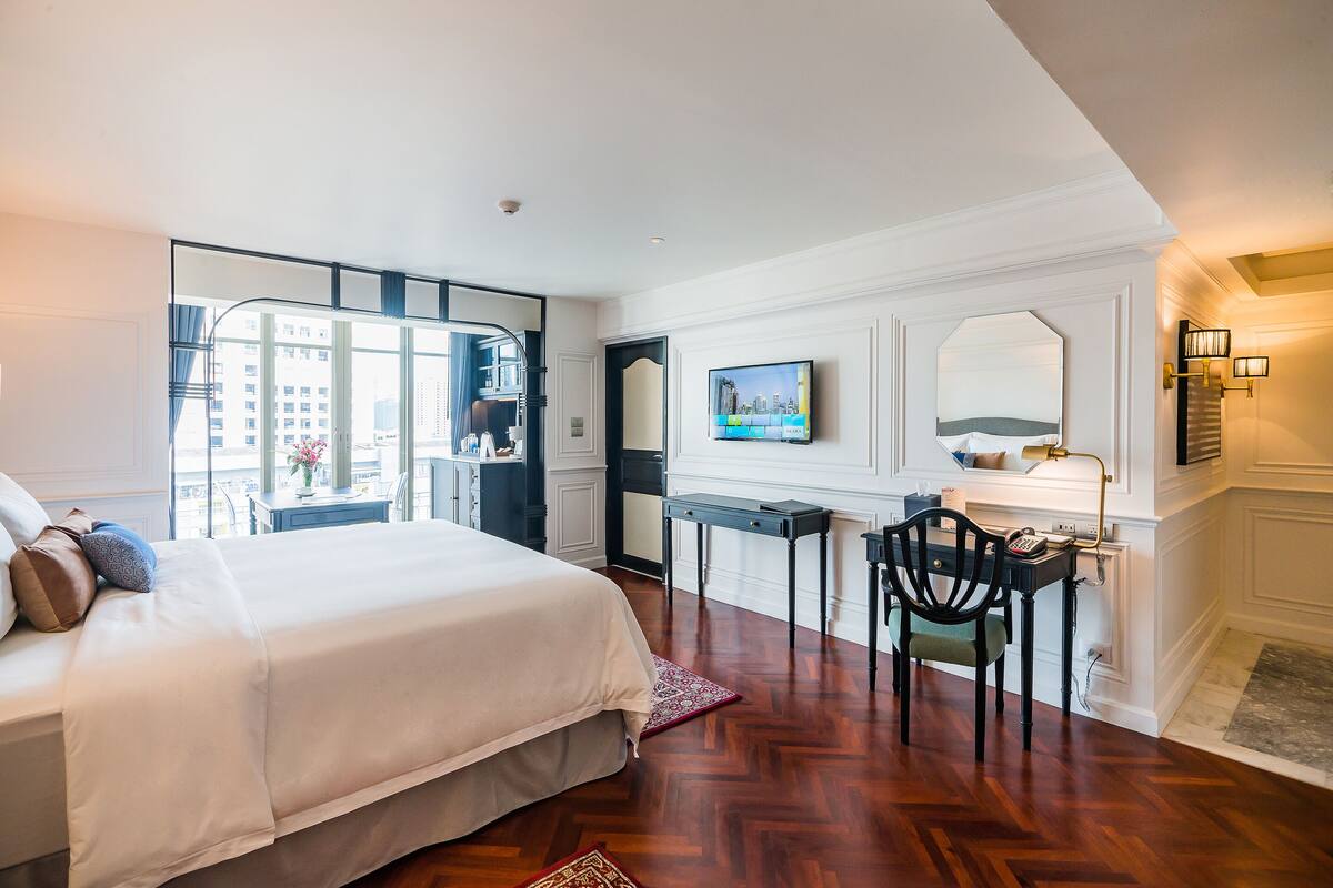A hotel room with a bed, a desk, a chair, a TV, and a view of the city through large windows.